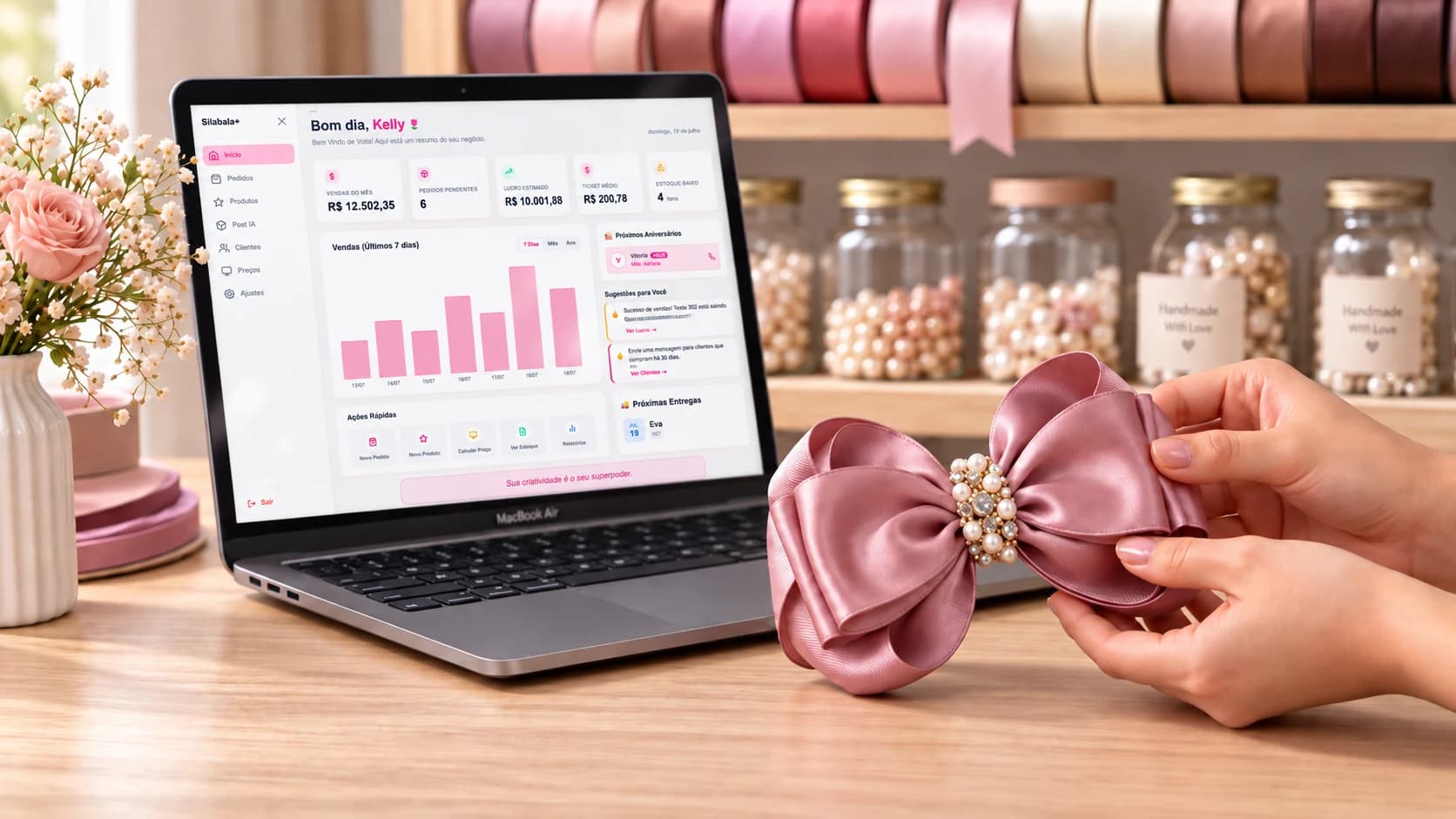 Notebook aberto no painel do Silabala+ mostrando vendas do mês e pedidos, ao lado de mãos montando um laço rosa com fitas e pérolas ao fundo